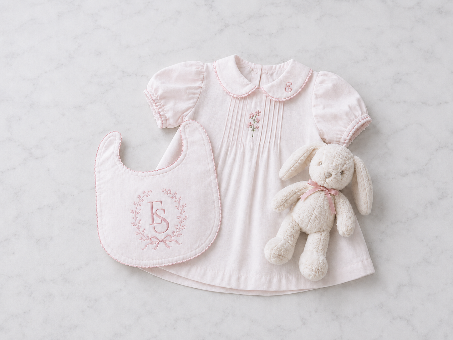 A pale pink baby dress with a monogrammed bib and a plush bunny, representing the Baby and Children collection